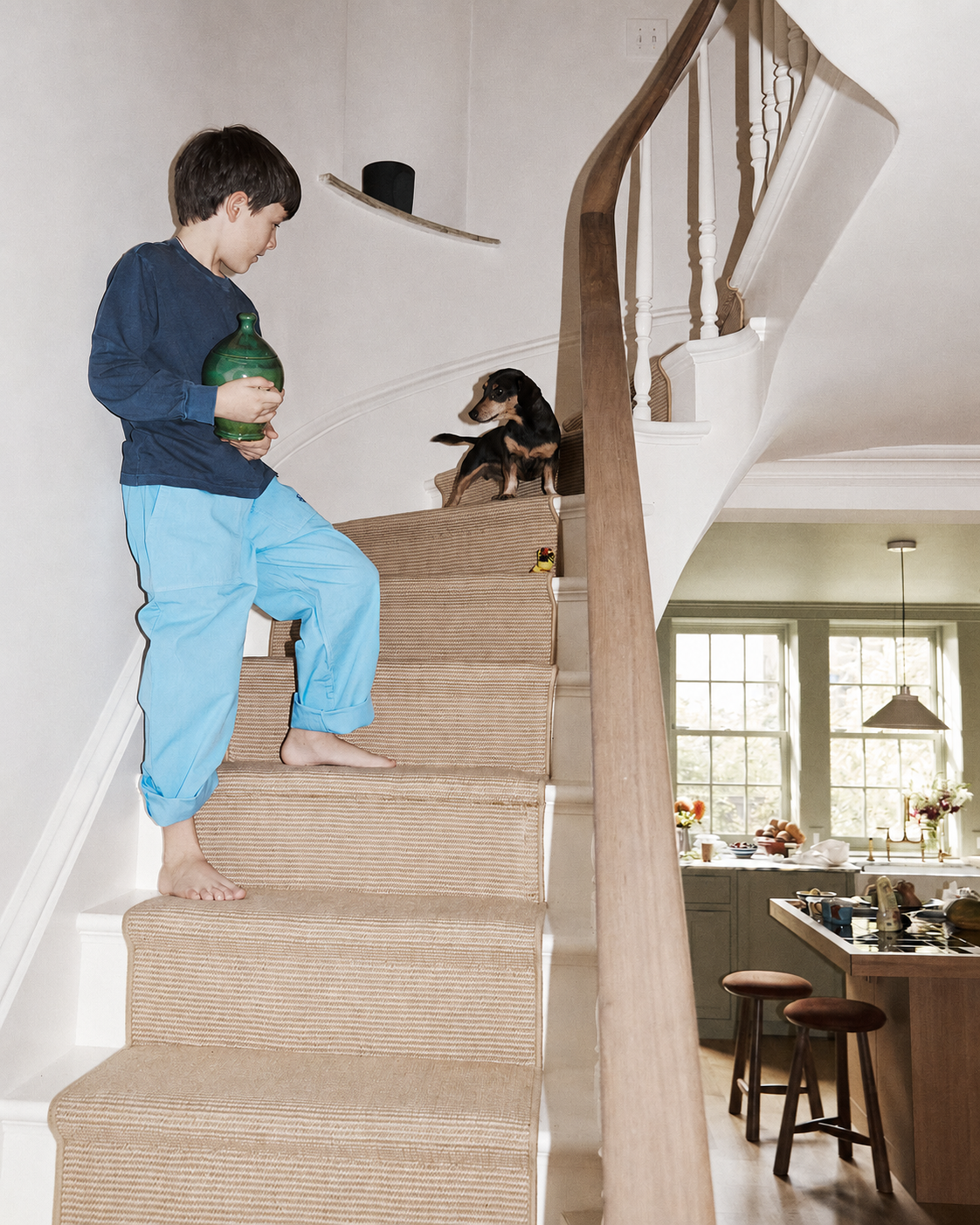 A boy in blue pajamas stands barefoot on the stairs, holding a Hugo Money Pot and looking at a small dog near the top of the steps. The kitchen is visible in the background.