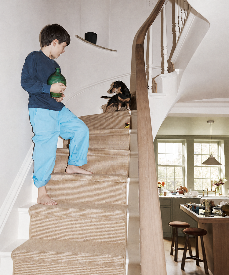A boy in blue pajamas stands barefoot on the stairs, holding a Hugo Money Pot and looking at a small dog near the top of the steps. The kitchen is visible in the background.