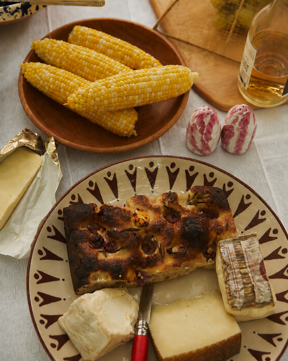 A table with corn on the cob, a bottle of liquor, Radicchio Salt and Pepper Shakers, and a plate with three types of cheese and a slice of bread.