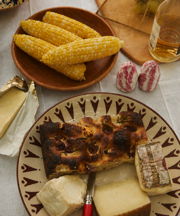 A table with corn on the cob, a bottle of liquor, Radicchio Salt and Pepper Shakers, and a plate with three types of cheese and a slice of bread.