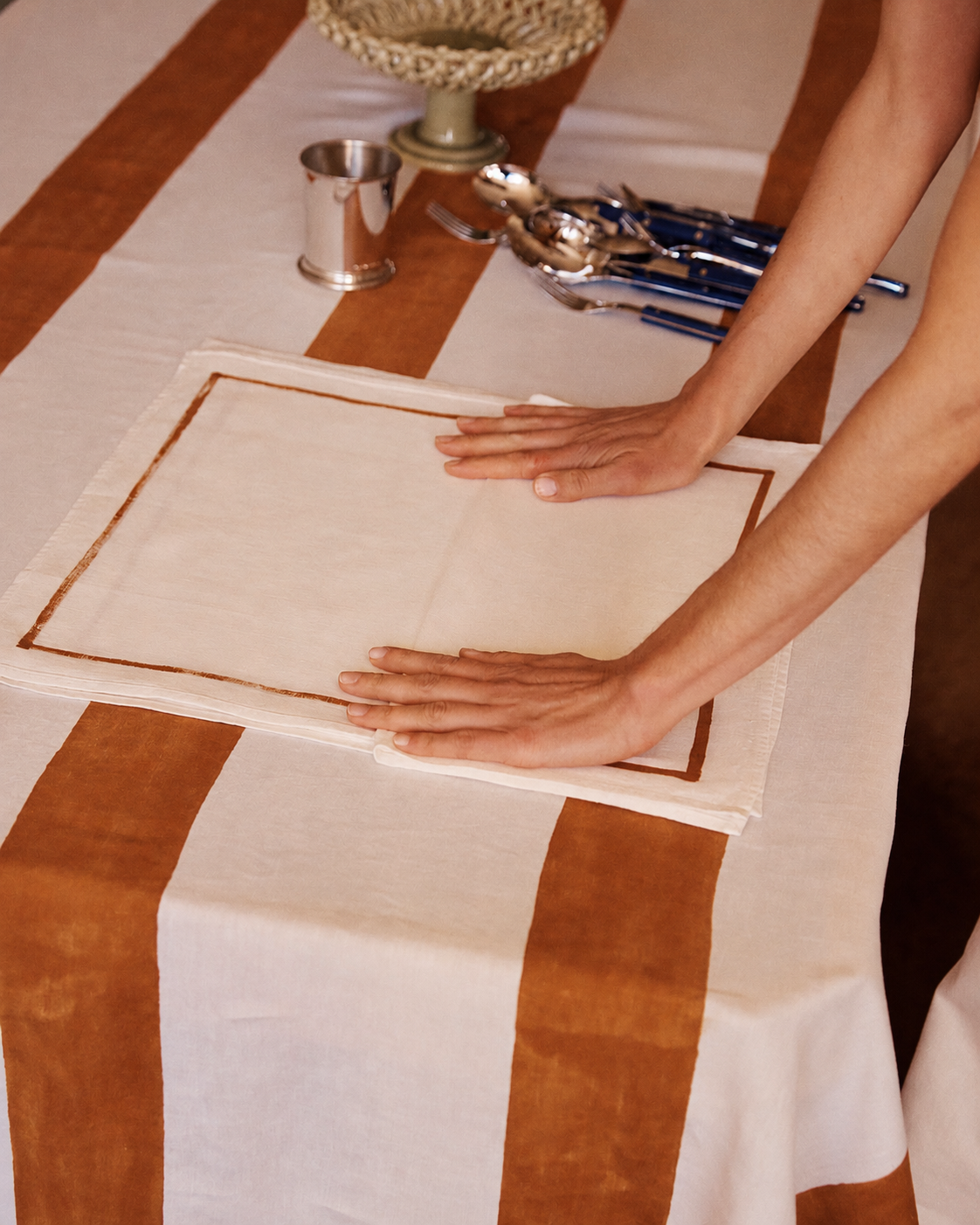 A person smooths a white placemat with brown trim on a table set with brown and white striped cloth; the Riga Napkin, Set of 6, handmade cutlery, cup, and bowl are also visible.
