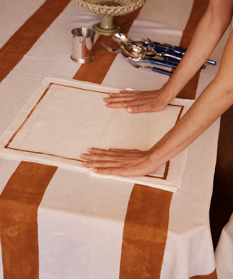 A person smooths a white placemat with brown trim on a table set with brown and white striped cloth; the Riga Napkin, Set of 6, handmade cutlery, cup, and bowl are also visible.