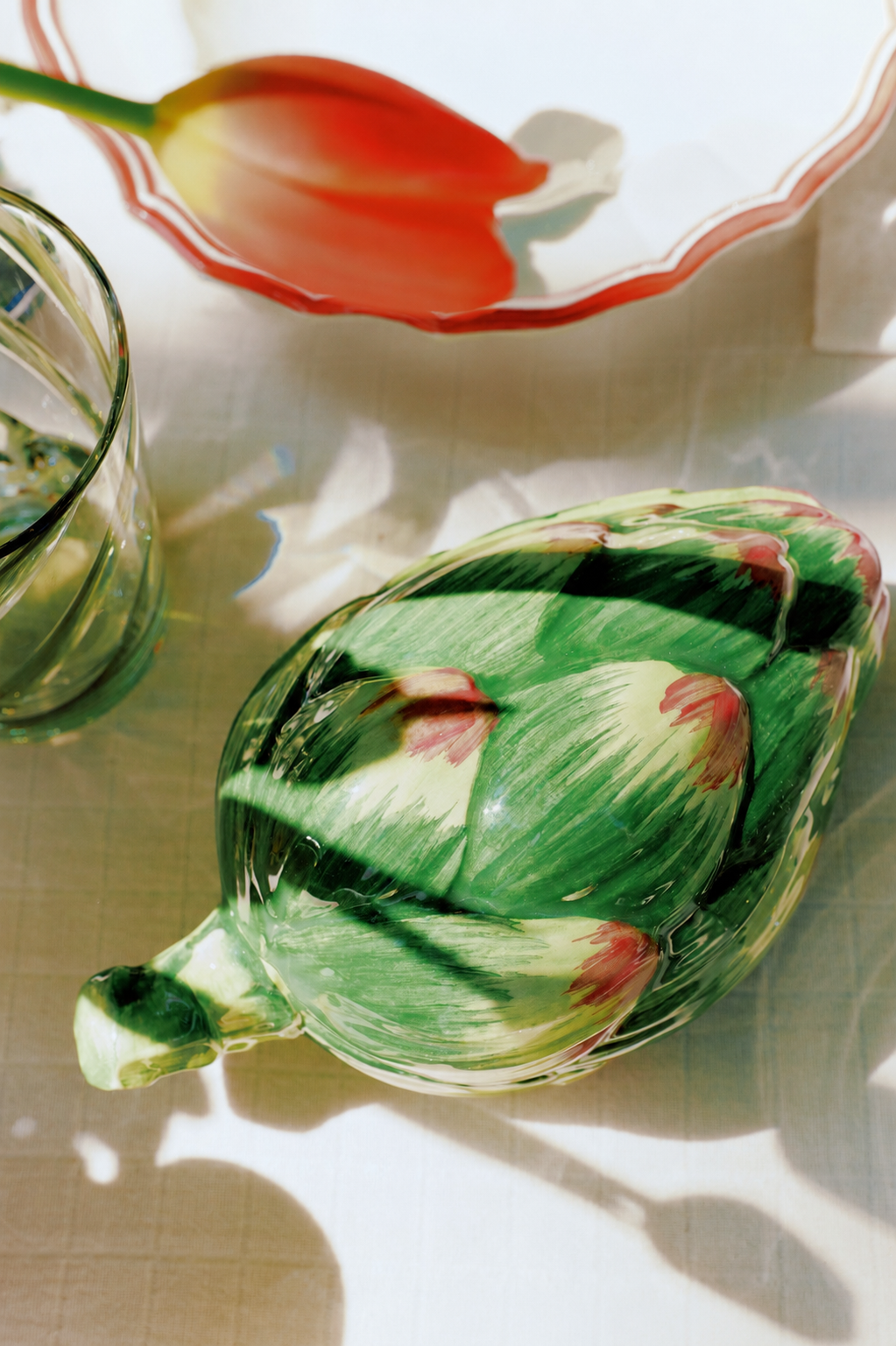 The Artichoke Sauce Bowl sits on a table beside a glass of water, with the shadow of a tulip dish in the background—an elegant touch of Italian kitchen tableware.