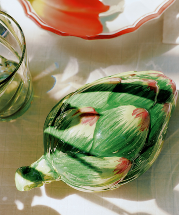 The Artichoke Sauce Bowl sits on a table beside a glass of water, with the shadow of a tulip dish in the background—an elegant touch of Italian kitchen tableware.