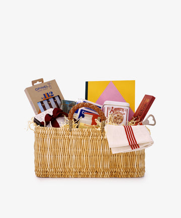 A Reed Basket with Leather Handles filled with packaged food items, a striped towel, books, and a heart-shaped wooden item, arranged against a plain white background.