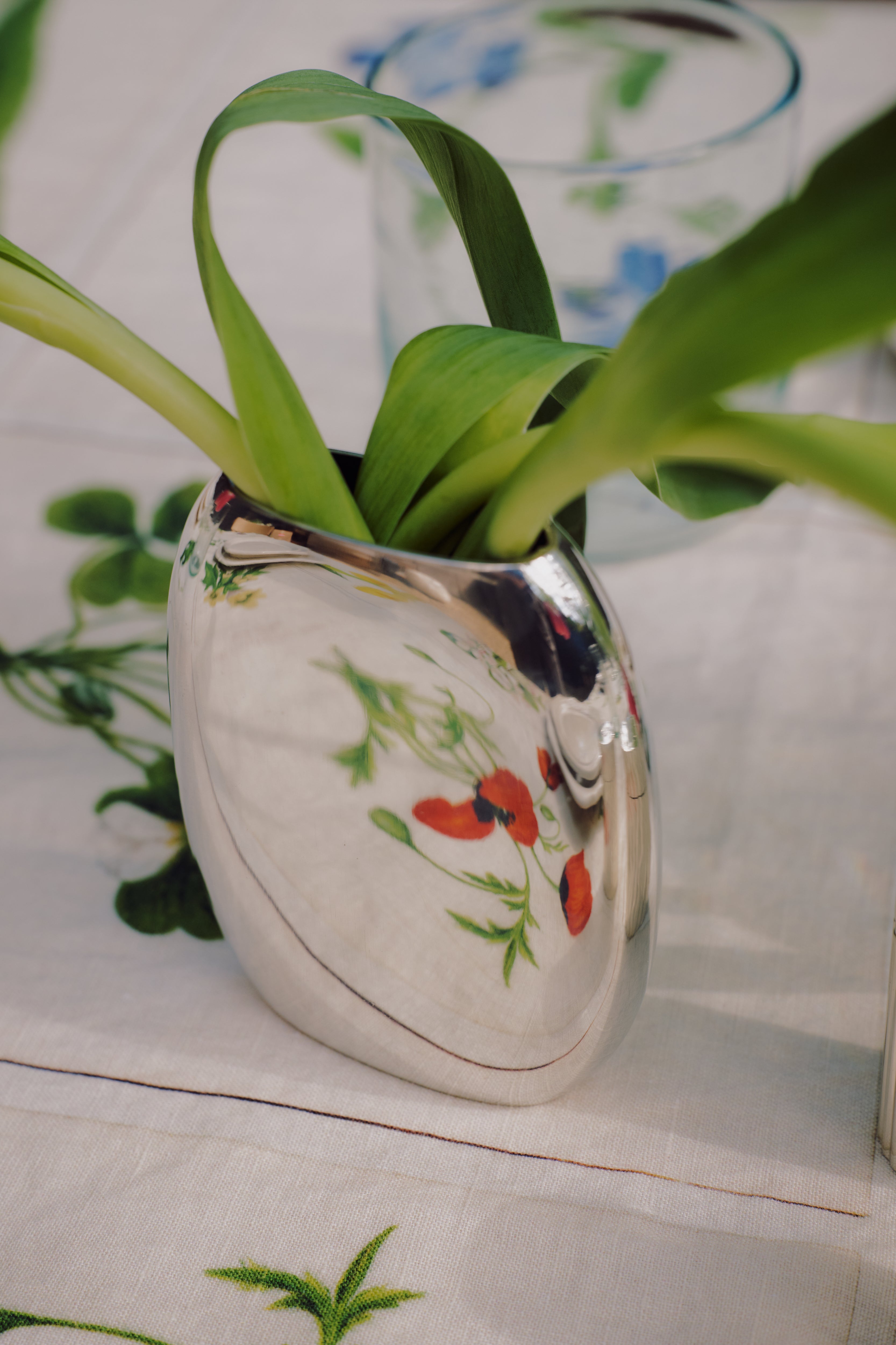 The Vera Oval Budvase, featuring a pewter finish and green leaves, sits on a white tablecloth adorned with colorful floral patterns.