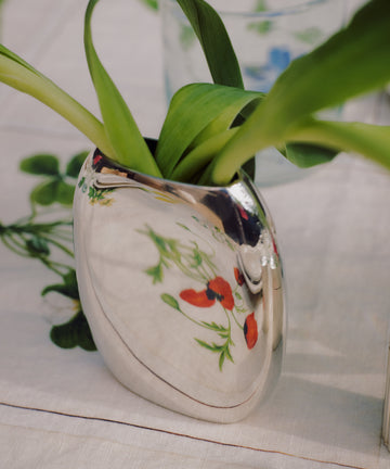 The Vera Oval Budvase, featuring a pewter finish and green leaves, sits on a white tablecloth adorned with colorful floral patterns.