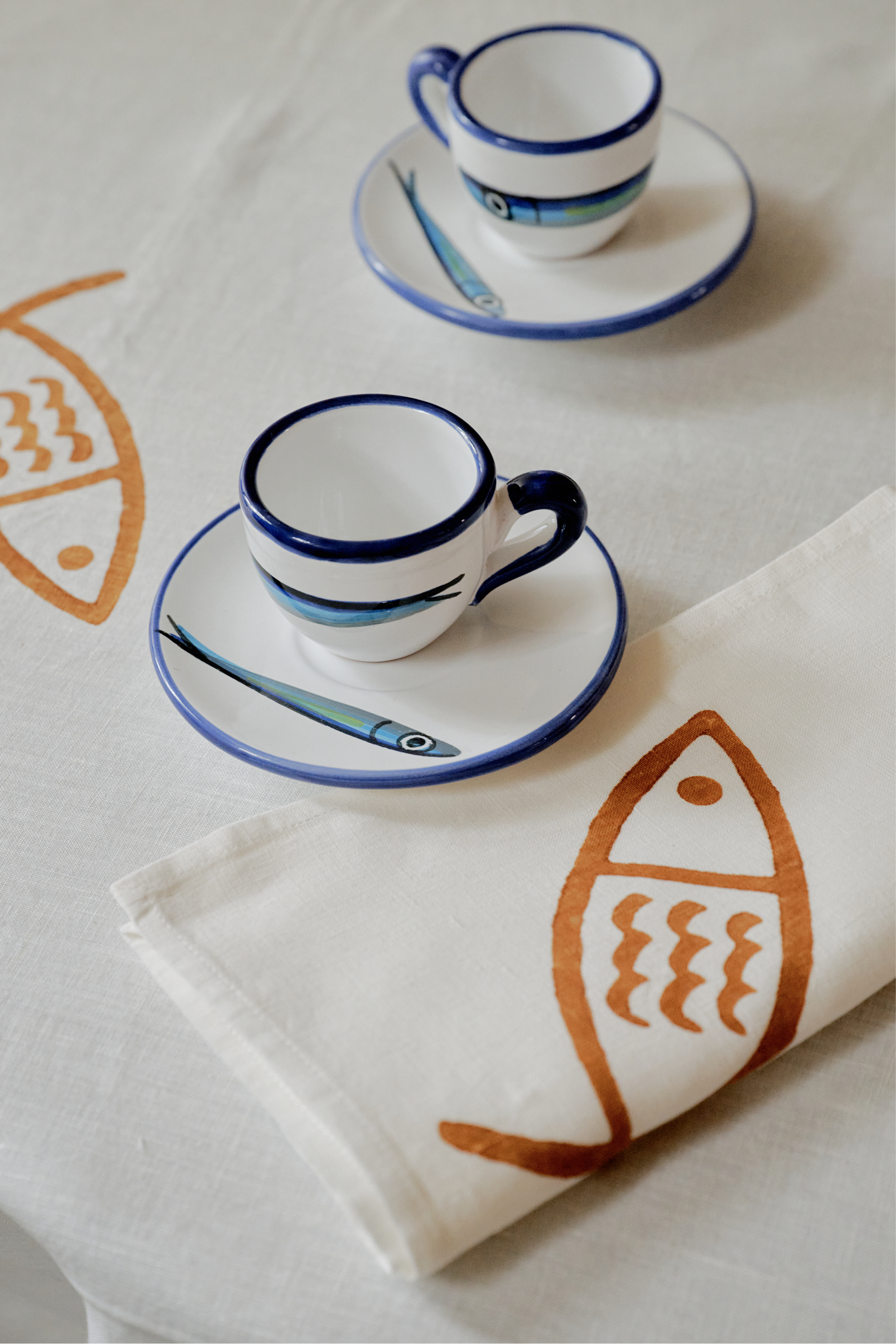 Two white espresso cups with blue rims and fish-adorned saucers sit on a table dressed in coastal linens and Pezza Napkins, Set of 6—hand block-printed napkins decorated with lively orange fish motifs.