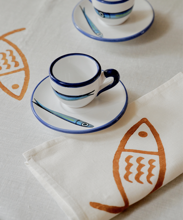 Two white espresso cups with blue rims and fish-adorned saucers sit on a table dressed in coastal linens and Pezza Napkins, Set of 6—hand block-printed napkins decorated with lively orange fish motifs.