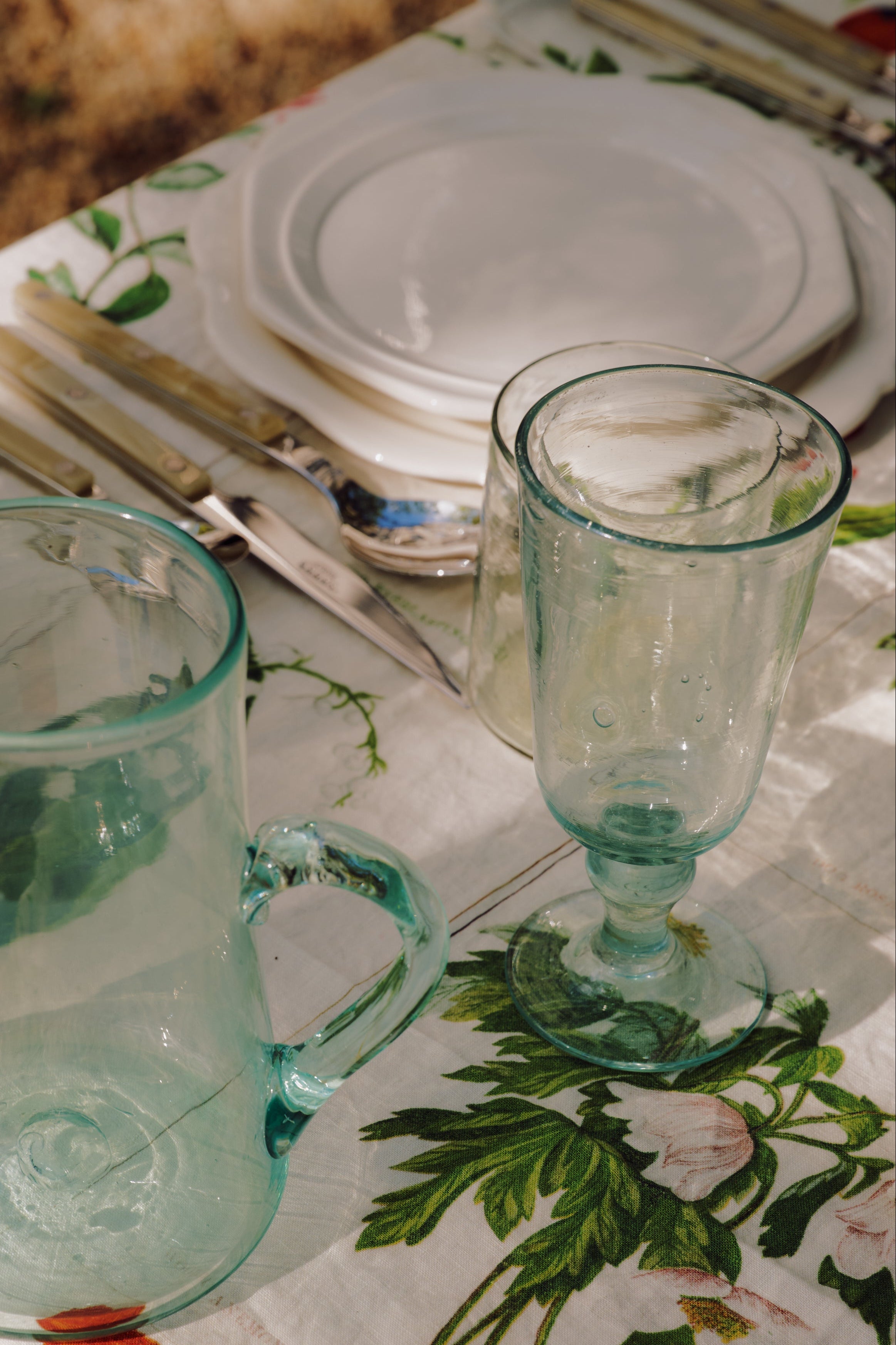 Elodie Stemmed Wine Glasses (set of 6), stacked white plates, and cutlery are arranged on a floral tablecloth in natural sunlight.