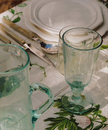 Elodie Stemmed Wine Glasses (set of 6), stacked white plates, and cutlery are arranged on a floral tablecloth in natural sunlight.