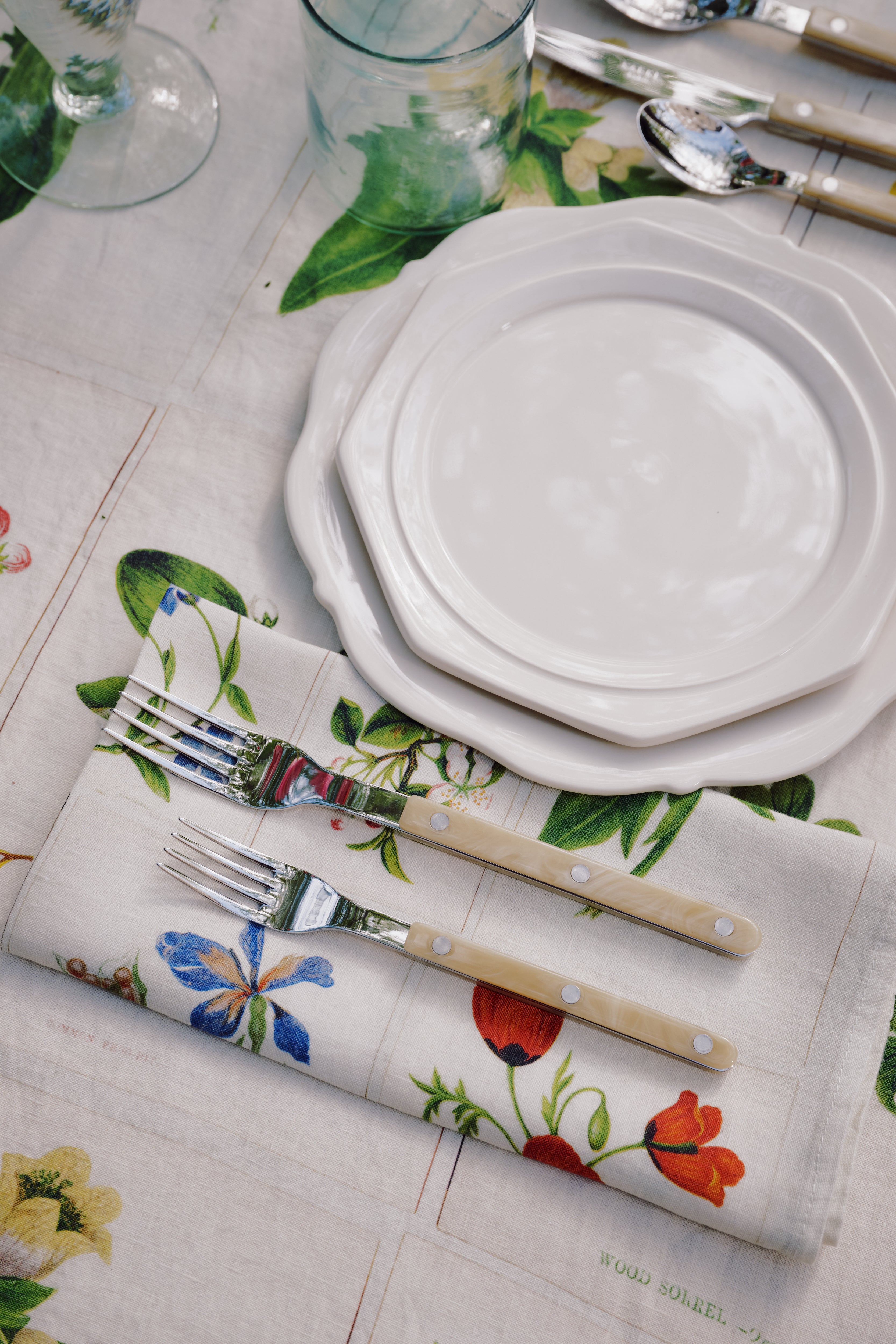 Two stacked Cirrus Scalloped Plates with cutlery are set on a floral napkin and matching tablecloth, surrounded by glasses and utensils, showcasing the beauty of classic creamware design.