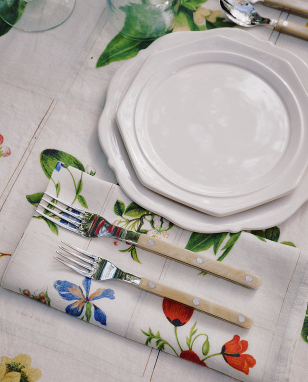 Two stacked Cirrus Scalloped Plates with cutlery are set on a floral napkin and matching tablecloth, surrounded by glasses and utensils, showcasing the beauty of classic creamware design.