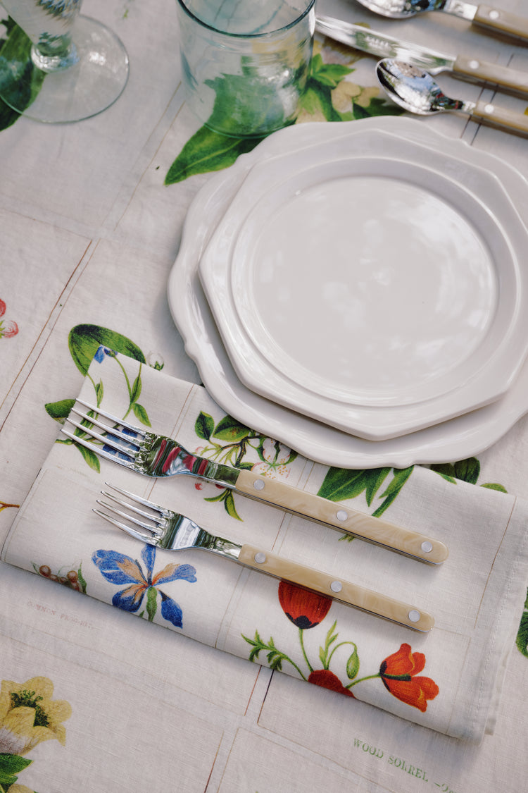 Two stacked Cirrus Scalloped Plates with cutlery are set on a floral napkin and matching tablecloth, surrounded by glasses and utensils, showcasing the beauty of classic creamware design.