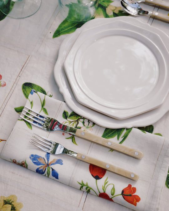 Two stacked Cirrus Scalloped Plates with cutlery are set on a floral napkin and matching tablecloth, surrounded by glasses and utensils, showcasing the beauty of classic creamware design.