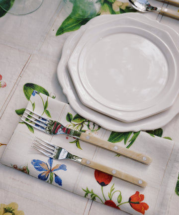 Two stacked Cirrus Scalloped Plates with cutlery are set on a floral napkin and matching tablecloth, surrounded by glasses and utensils, showcasing the beauty of classic creamware design.