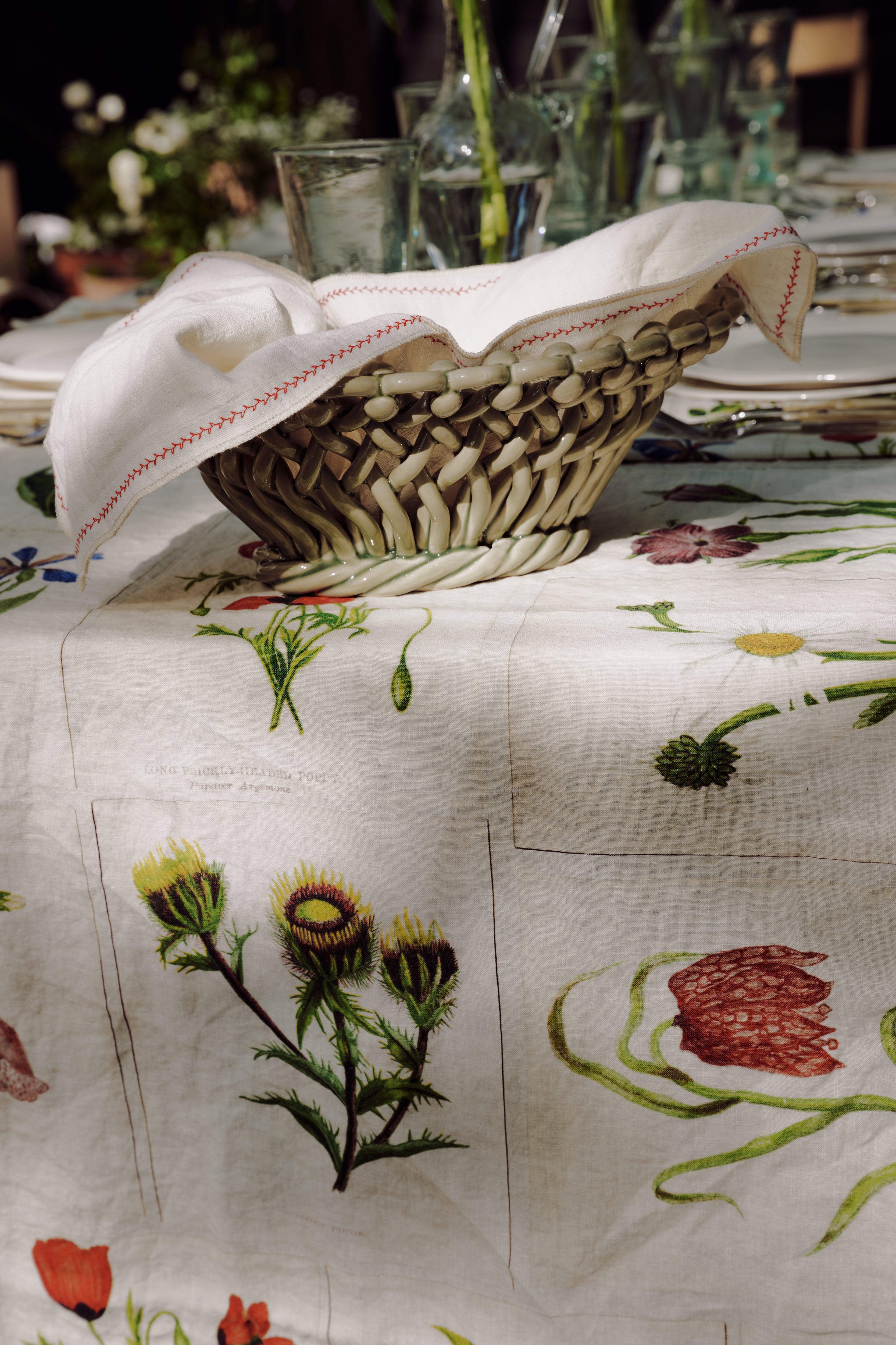 The Filial Bread Bento, Set of 2, features woven baskets with white cloths inspired by Japanese linens, displayed on a table with a botanical-patterned tablecloth and surrounded by elegant glassware and plates.