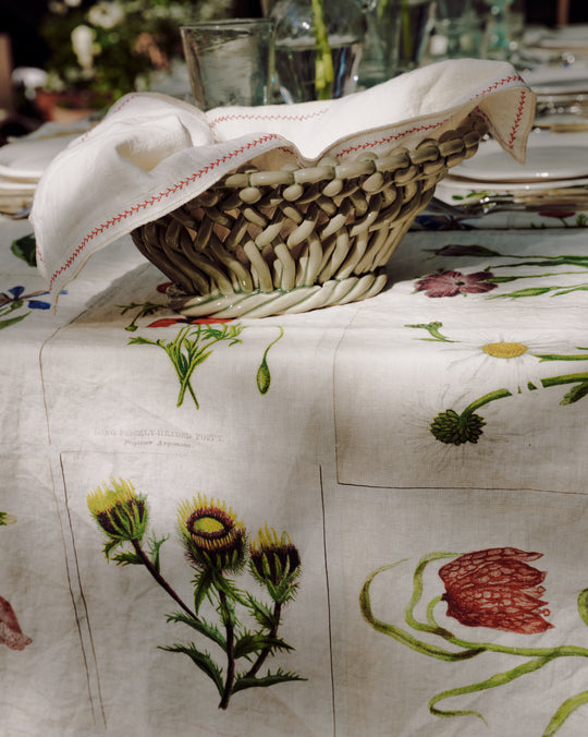 The Filial Bread Bento, Set of 2, features woven baskets with white cloths inspired by Japanese linens, displayed on a table with a botanical-patterned tablecloth and surrounded by elegant glassware and plates.