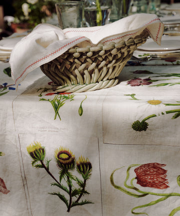 The Filial Bread Bento, Set of 2, features woven baskets with white cloths inspired by Japanese linens, displayed on a table with a botanical-patterned tablecloth and surrounded by elegant glassware and plates.