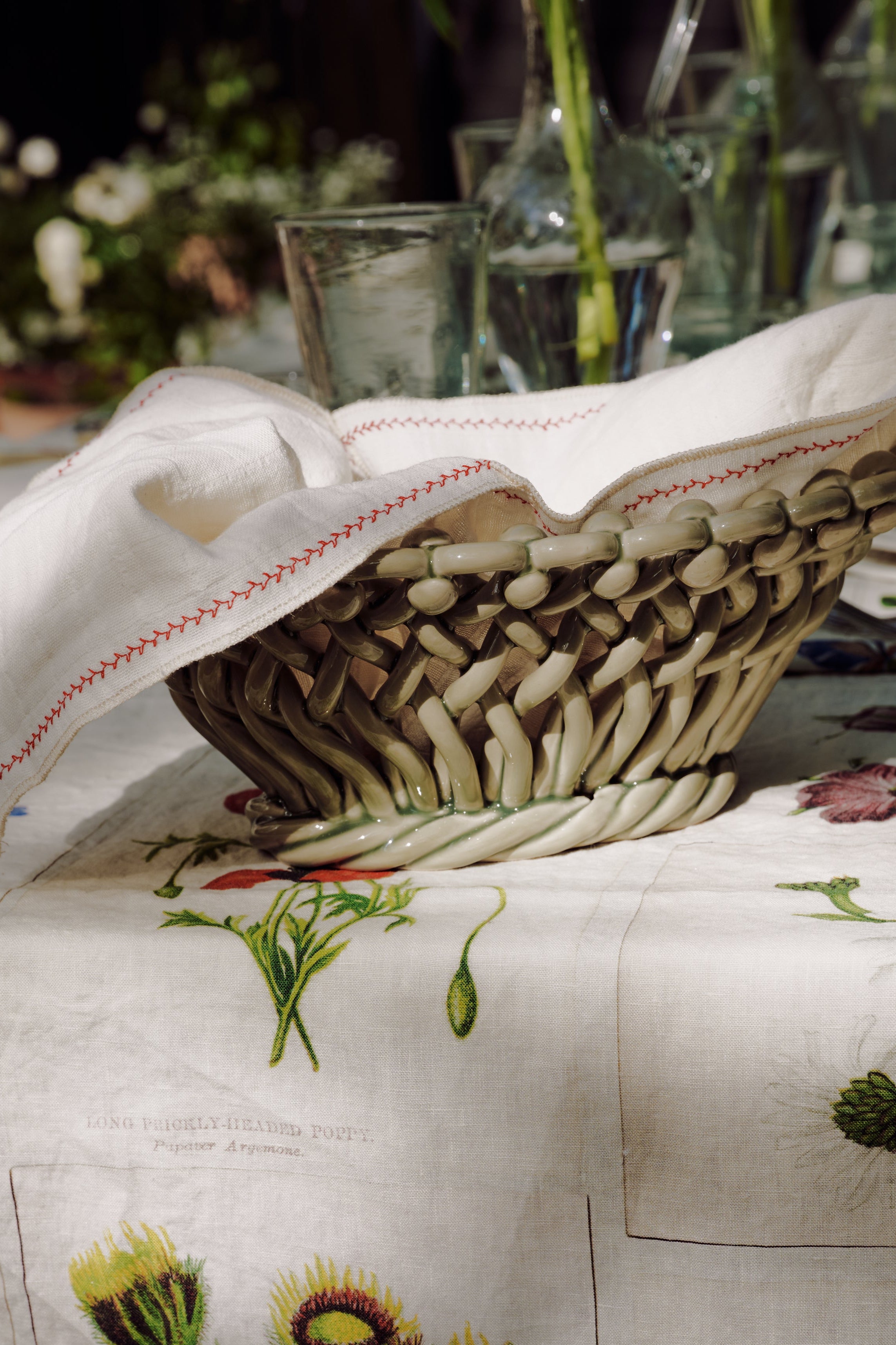 A woven basket with a white napkin sits on a botanical print tablecloth, surrounded by glassware and vases with green stems, evoking the relaxed elegance of the South of France. The scene is accented with Remi Decorative Flower Fruit Bowls, adding an artisanal touch to the inviting setting.