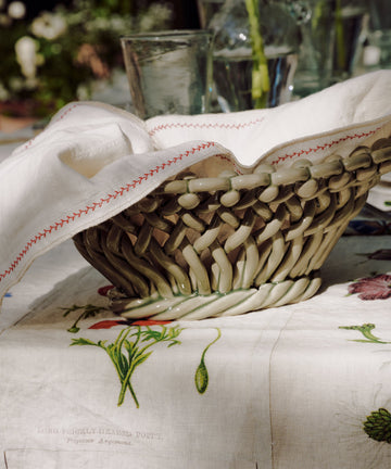 A woven basket with a white napkin sits on a botanical print tablecloth, surrounded by glassware and vases with green stems, evoking the relaxed elegance of the South of France. The scene is accented with Remi Decorative Flower Fruit Bowls, adding an artisanal touch to the inviting setting.