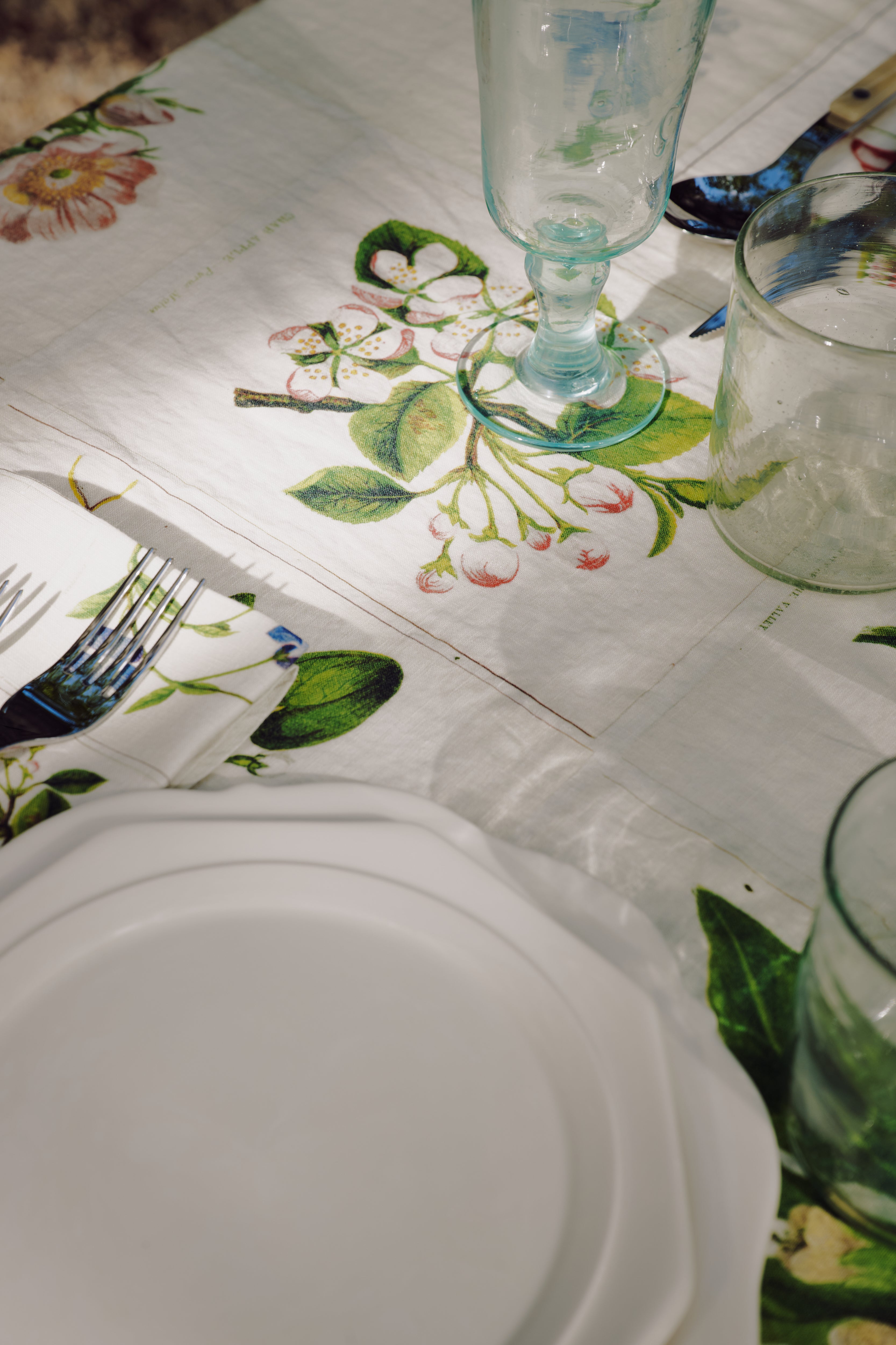 A white plate, glassware, and cutlery are set on a table covered with the John Derian Botanical Tiles Tablecloth, which features a delicate botanical design and is shown in natural light.