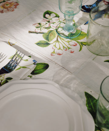 A white plate, glassware, and cutlery are set on a table covered with the John Derian Botanical Tiles Tablecloth, which features a delicate botanical design and is shown in natural light.