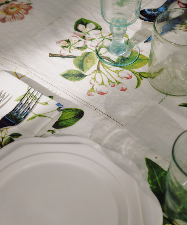 A white plate, glassware, and cutlery are set on a table covered with the John Derian Botanical Tiles Tablecloth, which features a delicate botanical design and is shown in natural light.