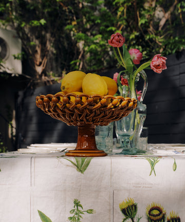 A white linen tablecloth with a vintage botanical pattern of flowers and leaves, styled with a woven fruit bowl, yellow lemons, and a glass vase of pink roses in an outdoor garden setting.
