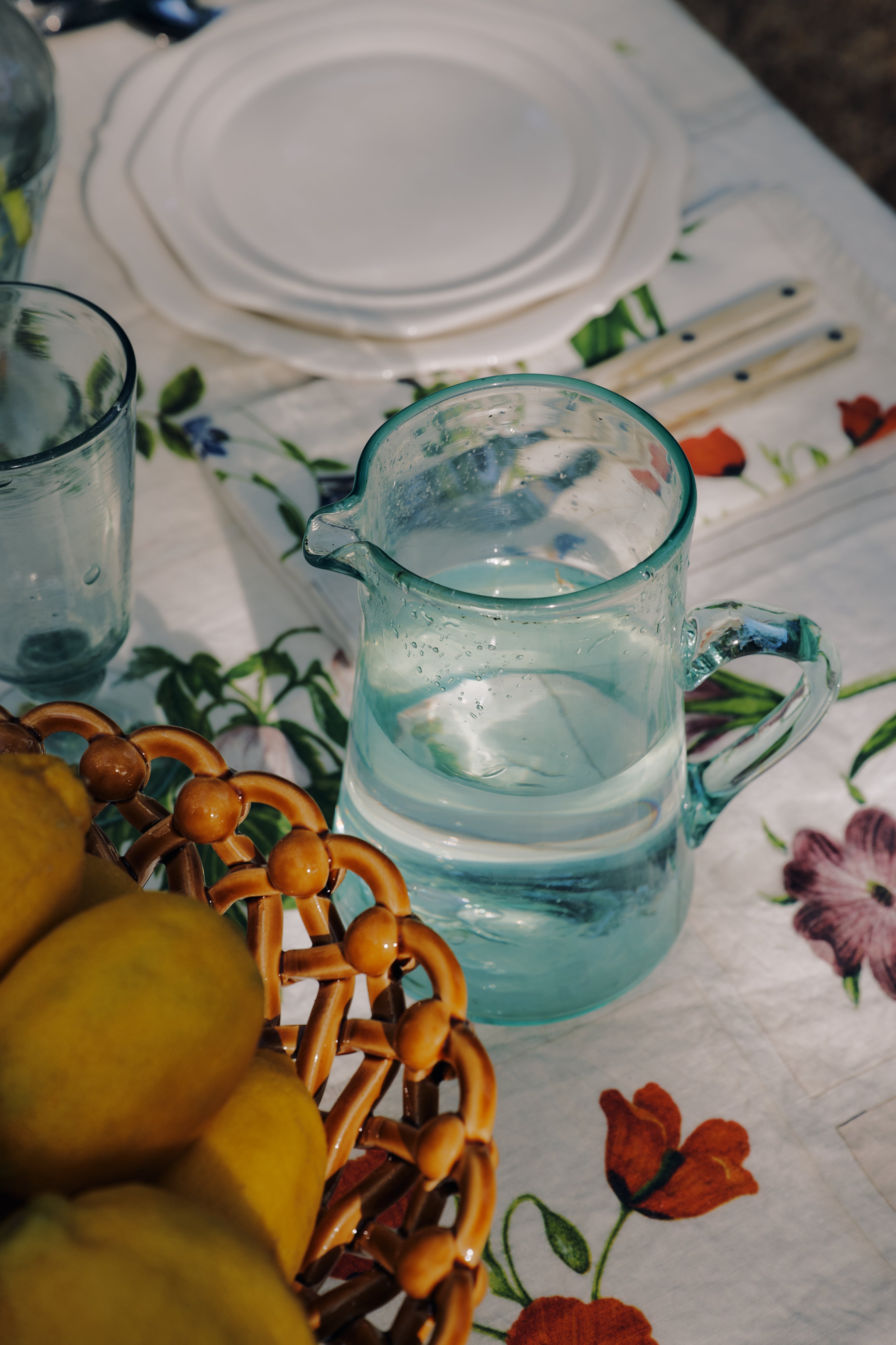 The Elodie Pitcher, a handblown clear glass piece from Damascus, sits on a floral tablecloth beside a basket of lemons, a glass, and stacked white plates with cutlery.