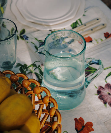 The Elodie Pitcher, a handblown clear glass piece from Damascus, sits on a floral tablecloth beside a basket of lemons, a glass, and stacked white plates with cutlery.