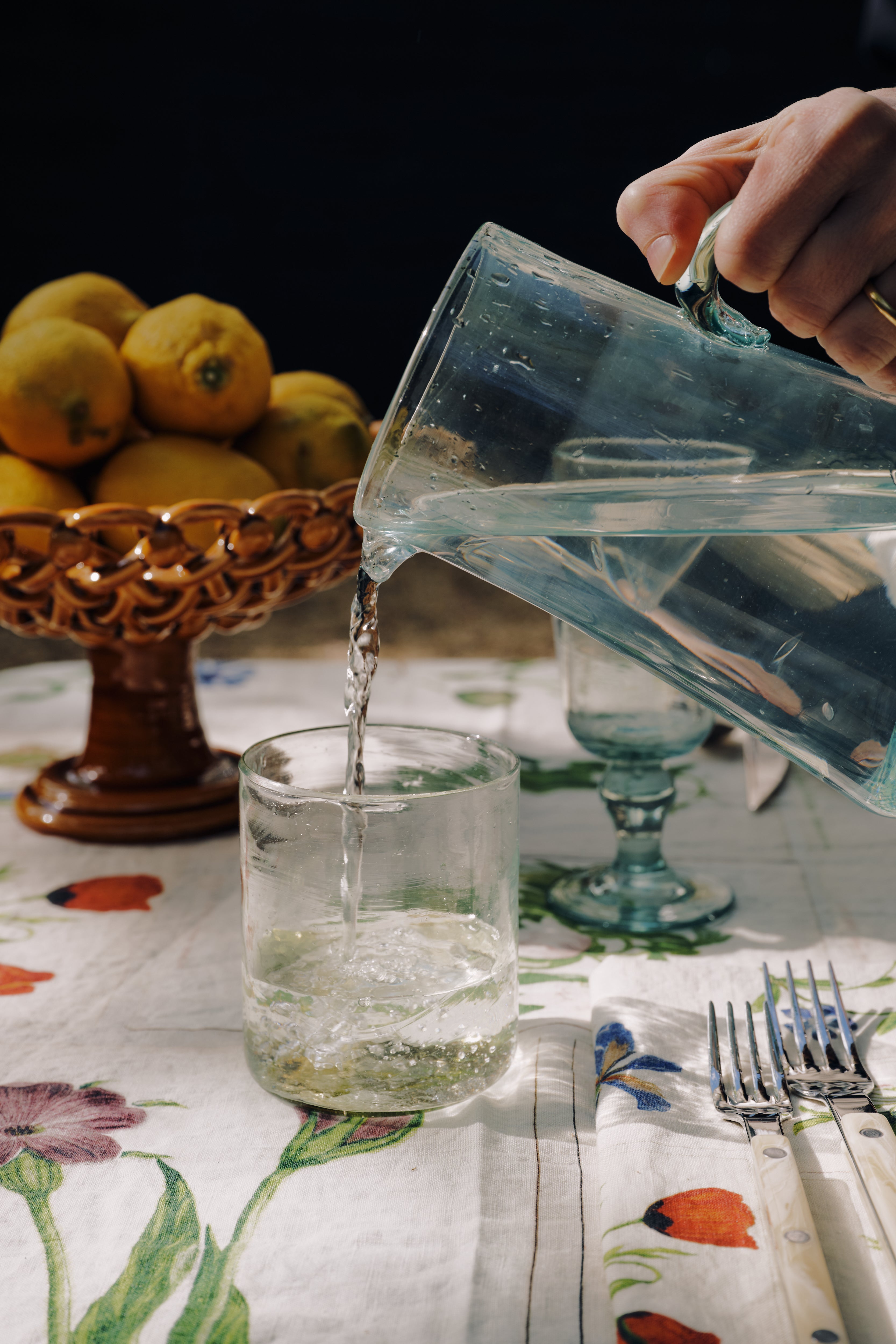 A hand pours water from a glass pitcher into an Elodie Straight Glass (set of 6) on a floral tablecloth, with a bowl of lemons and cutlery nearby.