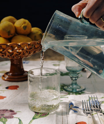 A hand pours water from a glass pitcher into an Elodie Straight Glass (set of 6) on a floral tablecloth, with a bowl of lemons and cutlery nearby.