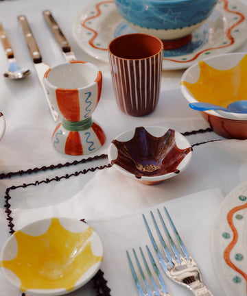 A table set with colorful ceramic plates, cups, cutlery, and unique kitchenware like the Stella Nesting Bowls and aperitivo snack bowls, all arranged on a white tablecloth with dark trim.