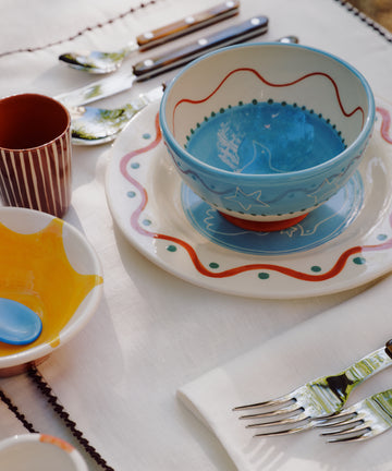 A set table features the Colombe Small Plate, a bowl stacked on a plate, a yellow dish with a spoon, silverware, and a cup, all arranged on a white placemat trimmed in brown for a colorful ceramic display.