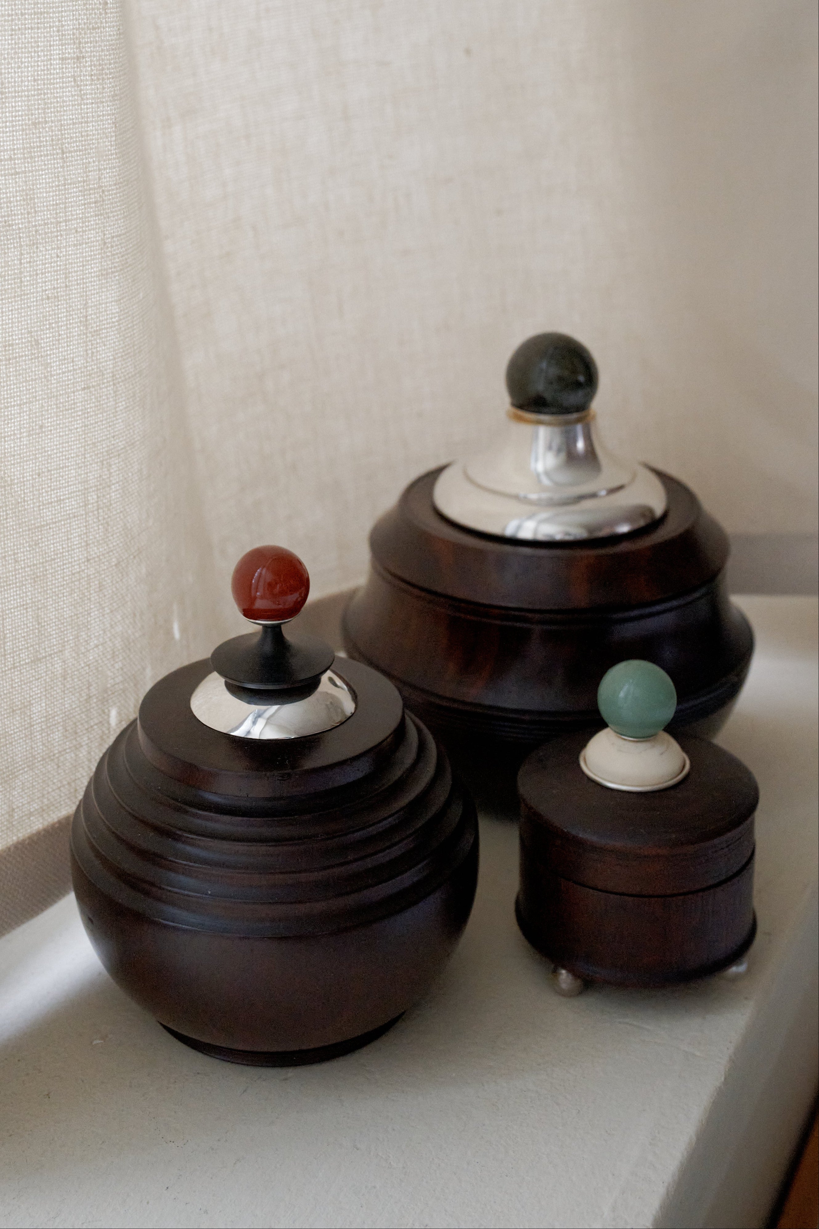 Three Curiosity Box round African wood boxes with metal lids and colorful knobs are displayed on a white surface next to a beige curtain.