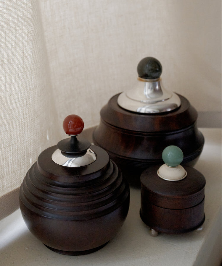 Three Curiosity Box round African wood boxes with metal lids and colorful knobs are displayed on a white surface next to a beige curtain.