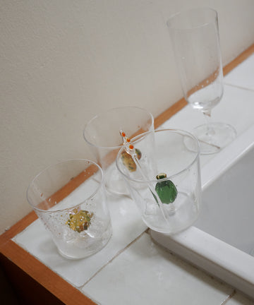 Four Lido Tumblers sit on a tiled countertop; three contain small figurines—a goldfish, frog, and turtle—while the fourth is a plain stemmed glass.