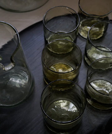 Six Elodie Straight Glasses, set of 6, filled with light liquid rest on a dark tray beside a glass pitcher, highlighting the elegance of recycled glass craftsmanship.