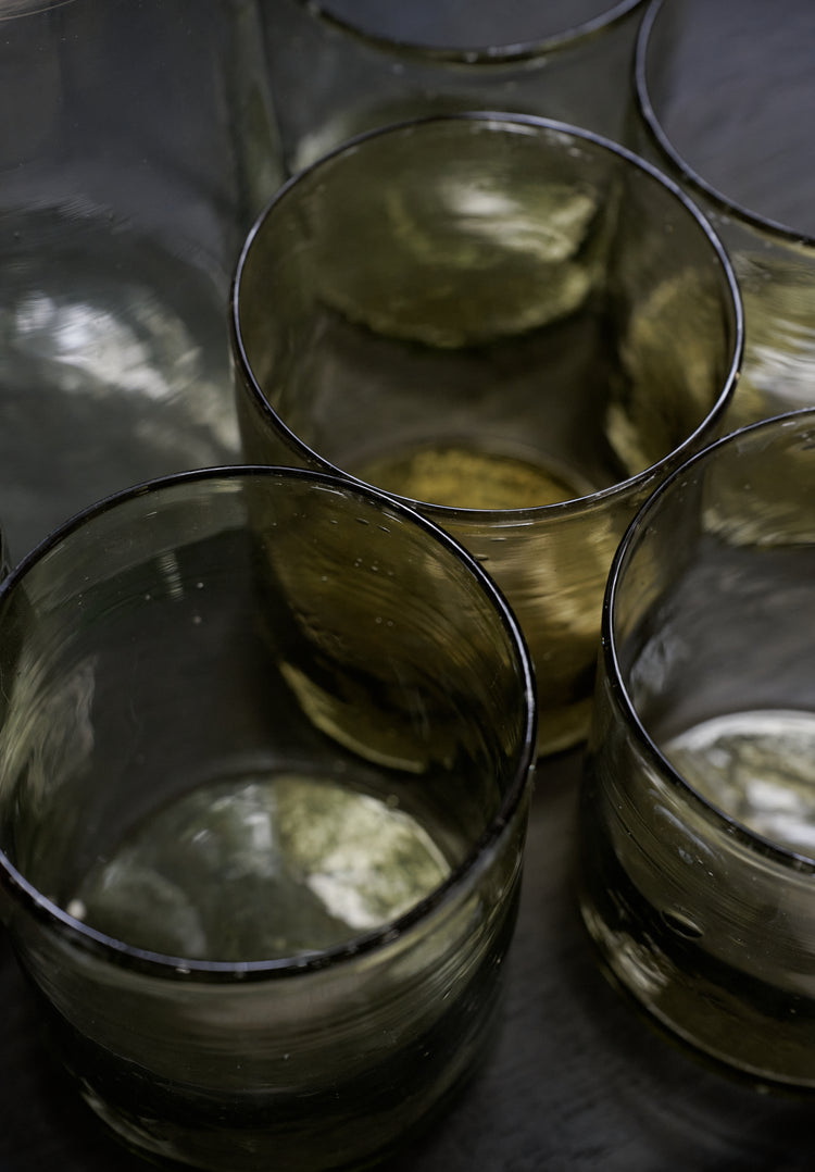A set of six Elodie Straight Glass tumblers, made from recycled translucent glass, are grouped together on a dark surface with light reflecting off their surfaces.