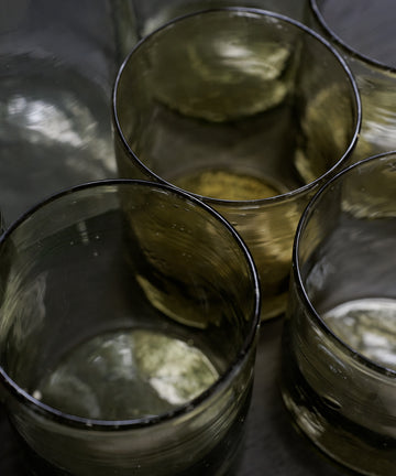 A set of six Elodie Straight Glass tumblers, made from recycled translucent glass, are grouped together on a dark surface with light reflecting off their surfaces.