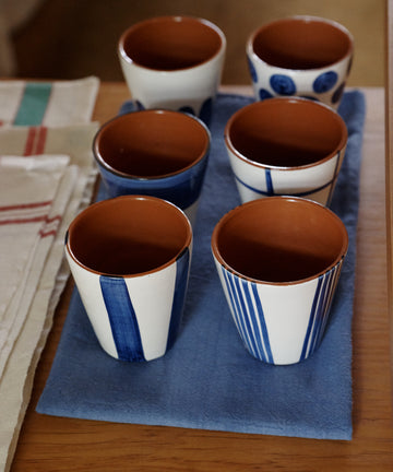 The Lula Cup, Set of 6, features blue and white ceramic cups crafted in a handmade pottery style. They are arranged on a blue cloth atop a wooden surface next to folded napkins.