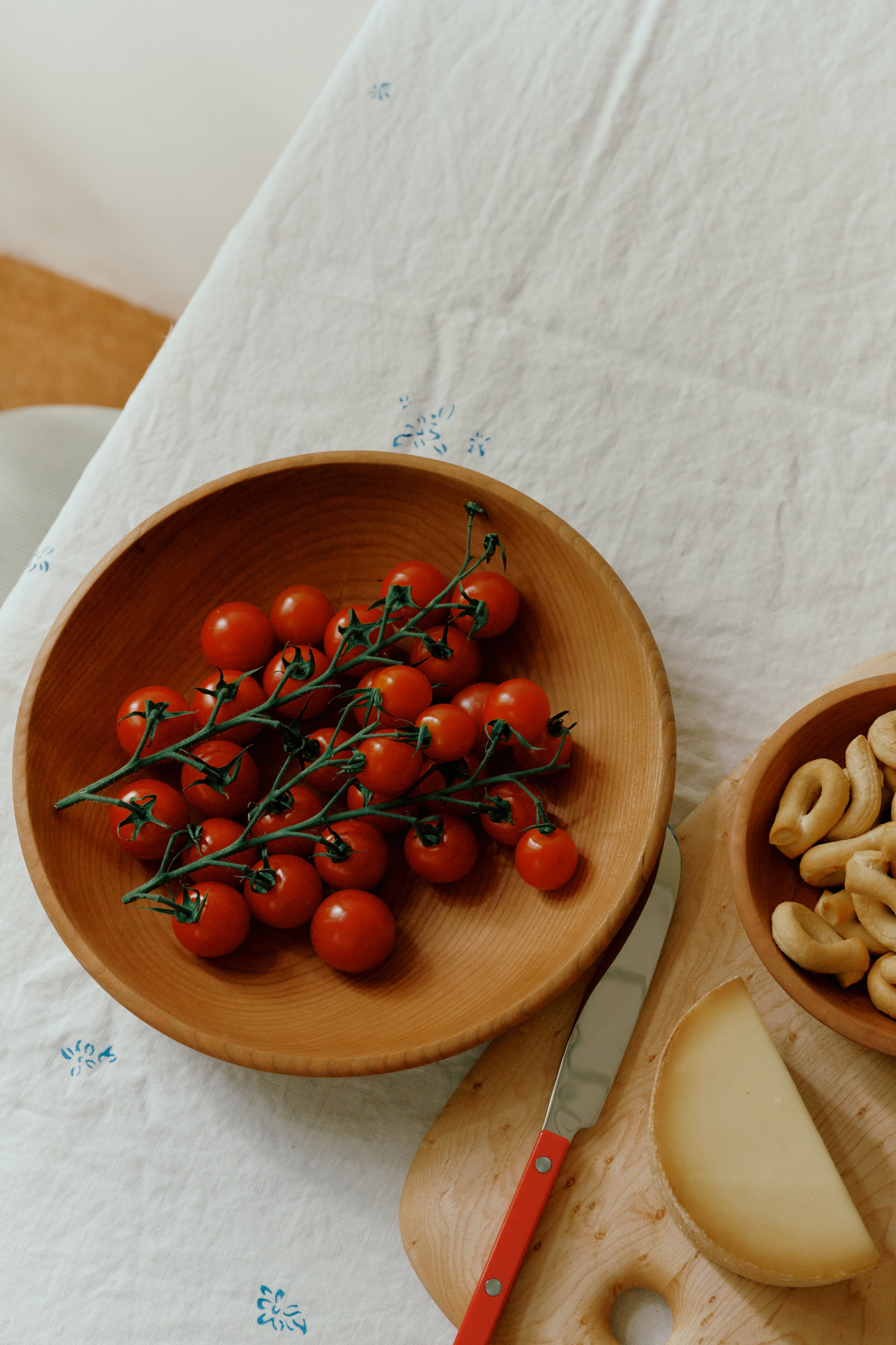 Wooden bowl with cherry tomatoes on a tablecloth with a knife and cheese.