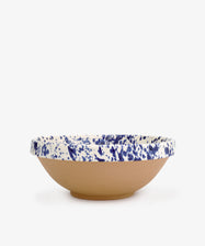 The Rita Bowl Splatter features a shallow, round design with a natural terracotta base and a white rim accented by blue splatter patterns, shown against a plain white background.