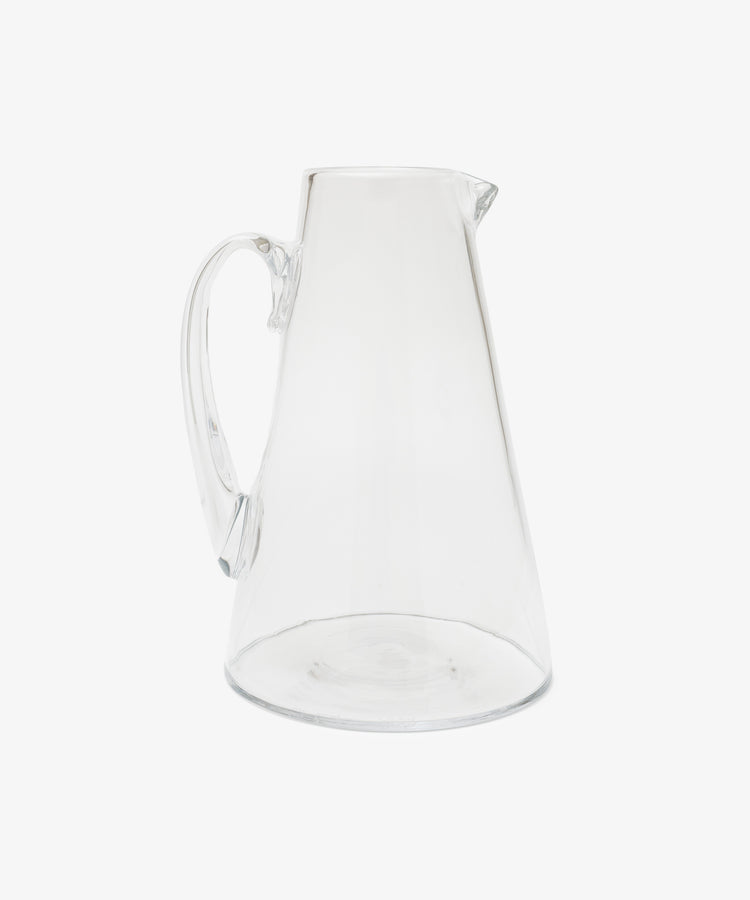 The Gemma Jug—a handmade, clear glass pitcher with a wide base, narrow top, curved handle, and spout—crafted in the South of France and inspired by oversized jugs, displayed on a plain white background.