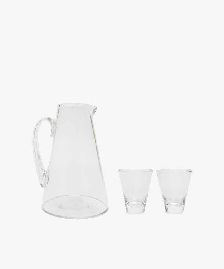 The Gemma Jug, a clear glass pitcher with a handle inspired by French handmade tradition, sits beside two empty clear glasses—all displaying crystal transparency against a plain white background.