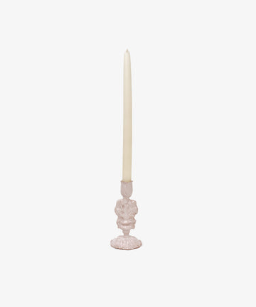 A tall, unlit white taper candle sits in the sculptural Jarra De Flores Candleholder, which features a decorative light pink glaze, set against a plain white background.