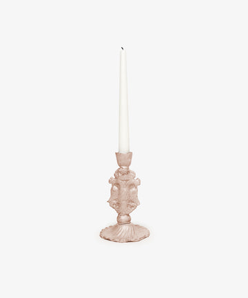 The Aguila Bicefala Candleholder, an ornate hand-moulded piece in light pink, displays a tall unlit white taper candle against a plain white background, emphasizing its sculptural elegance.