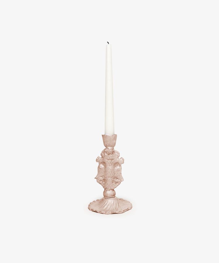 The Aguila Bicefala Candleholder, an ornate hand-moulded piece in light pink, displays a tall unlit white taper candle against a plain white background, emphasizing its sculptural elegance.