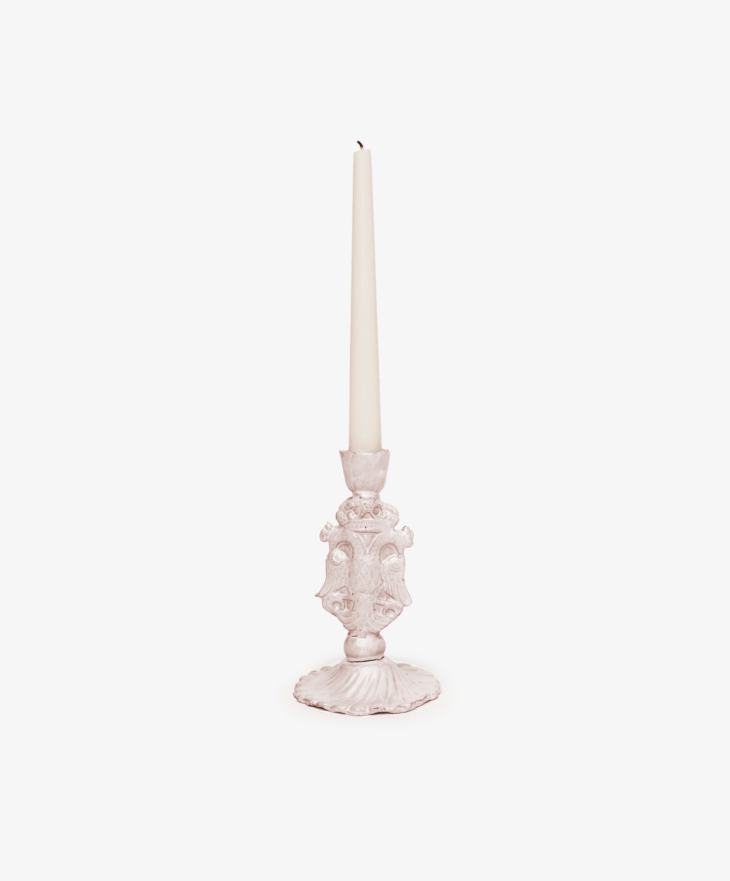 A white taper candle rests in the Aguila Bicefala Candleholder, an ornate, hand-moulded light pink piece, set against a plain white background.